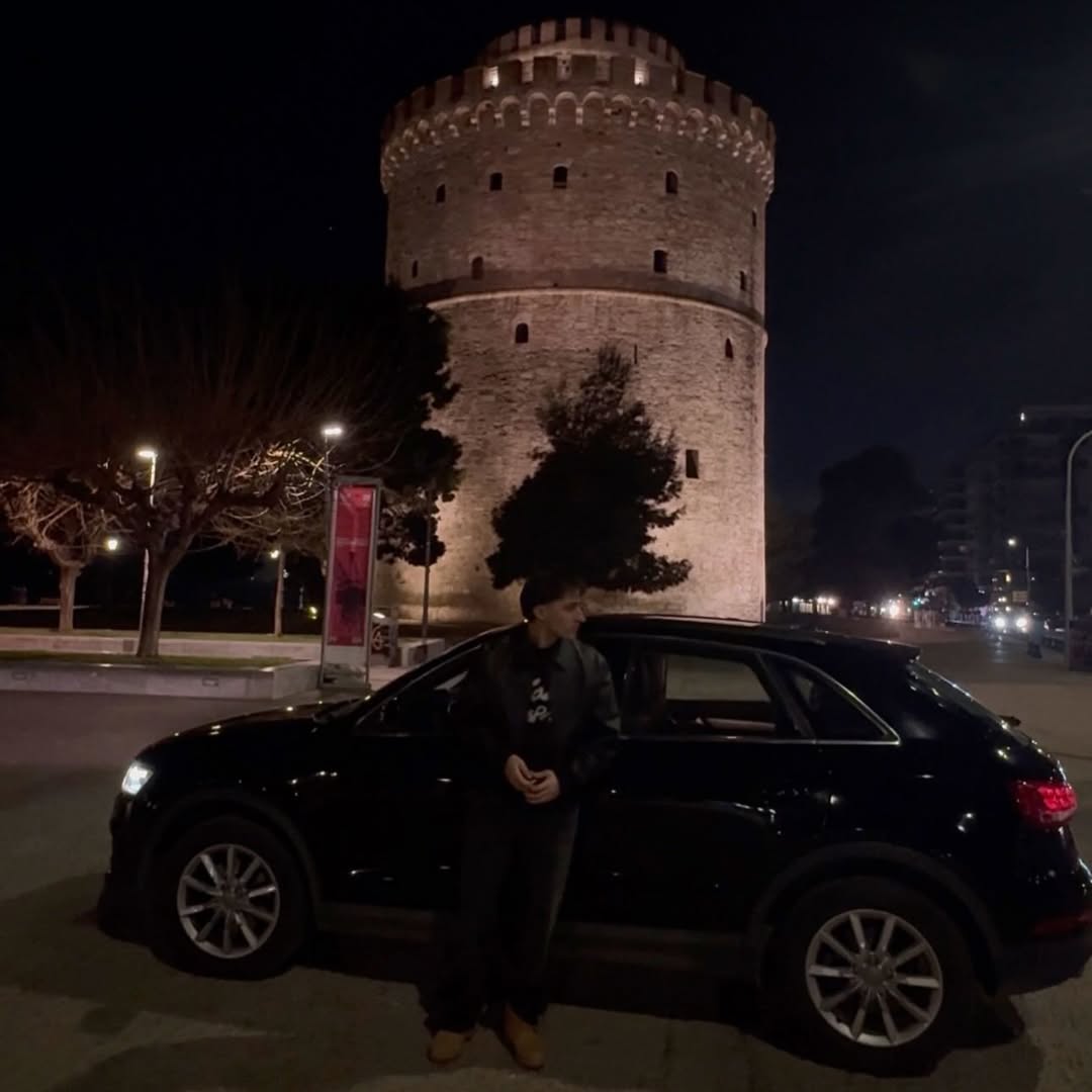 Standing next to a black Audi in front of the White Tower of Thessaloniki at night