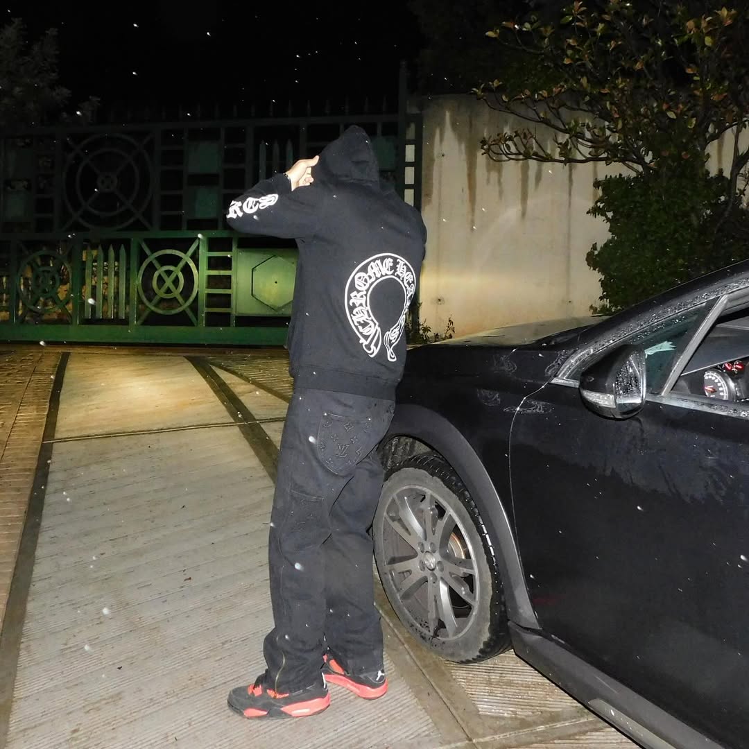 Back view in Chrome Hearts hoodie and Louis Vuitton jeans next to a black Maserati at night